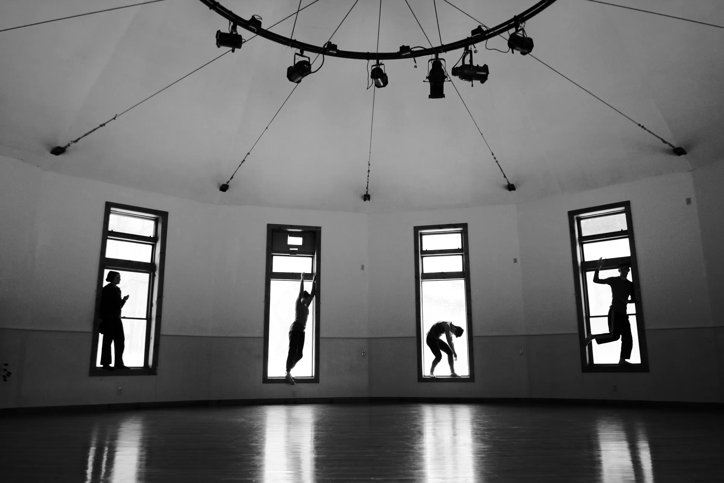 Four performers create distinct poses in the windows of a circular black-and-white rehearsal space.