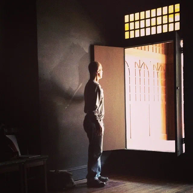 A workshop participant stands in a dark rehearsal room facing an open door filled with sunlight.