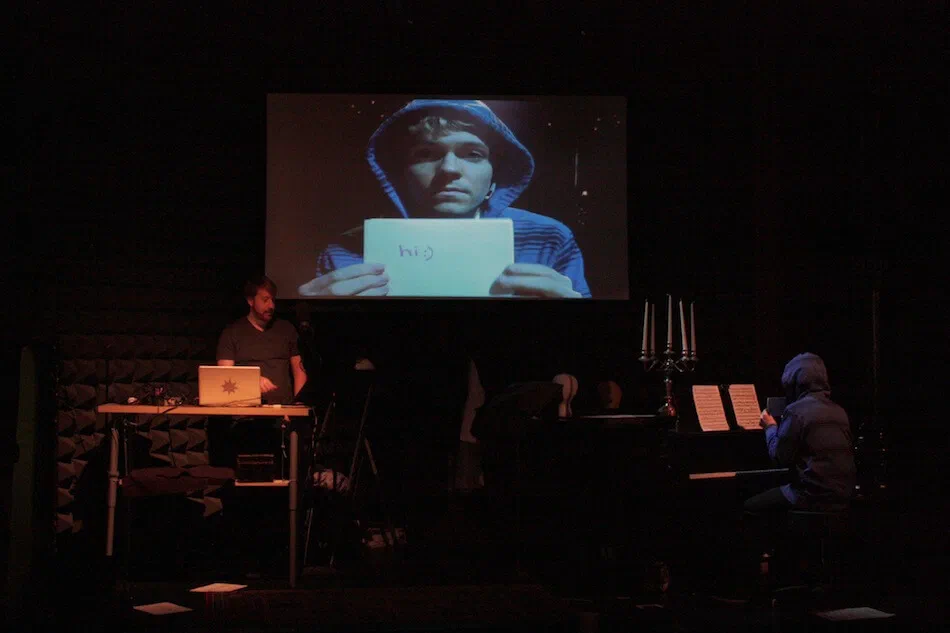 Musicians perform below a projection of a face holding a handwritten card.