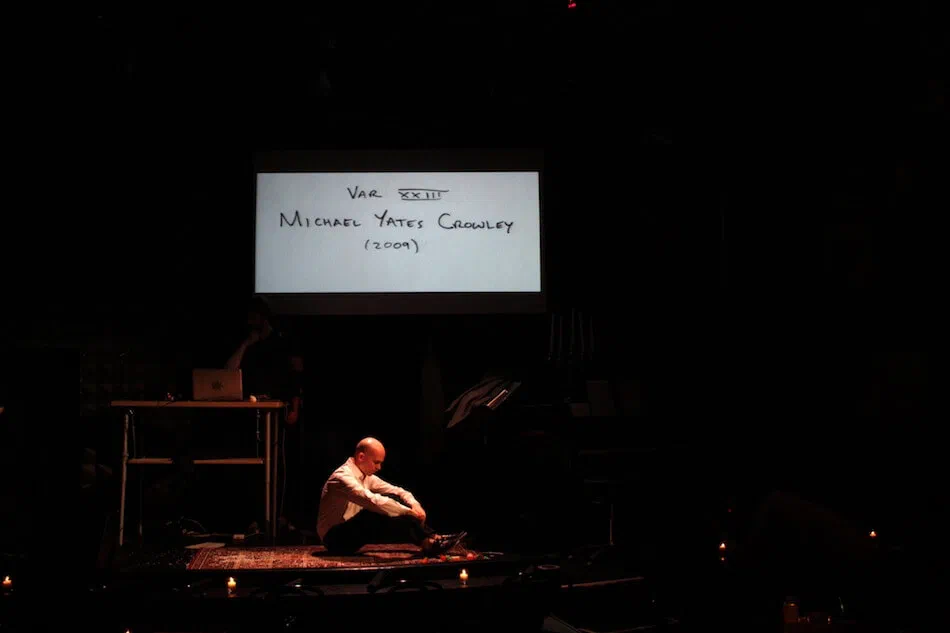 A performer sits on the floor beneath a screen displaying handwritten text.