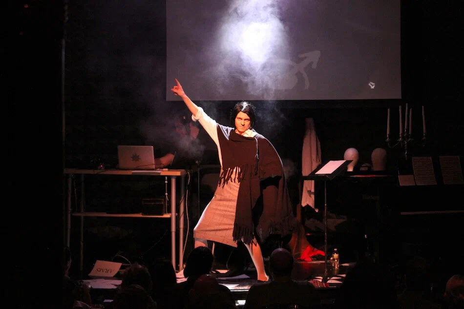 A performer raises one arm through a cloud of smoke on a dark stage.