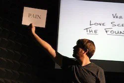A performer raises a card reading PAIN beside a screen of handwritten text.