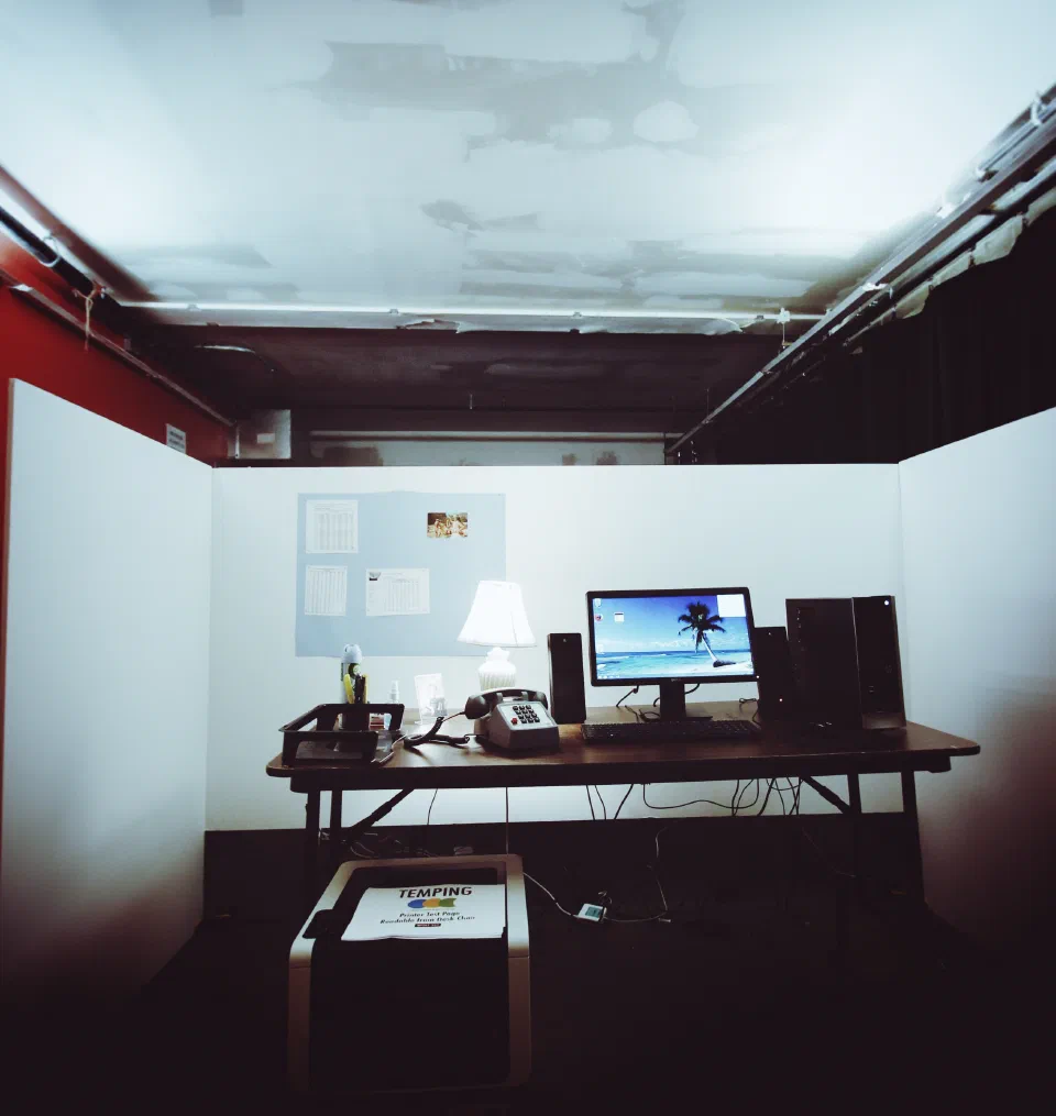 A temporary office installation contains a desk, computer, telephone, lamp, printer, and paperwork.