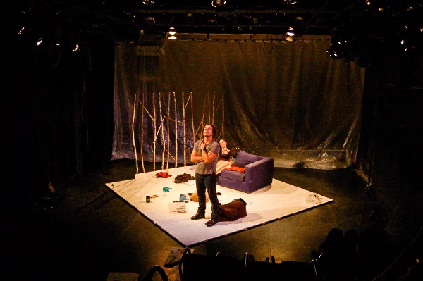 A performer stands amid scattered objects, a couch, and a row of slender bare branches.