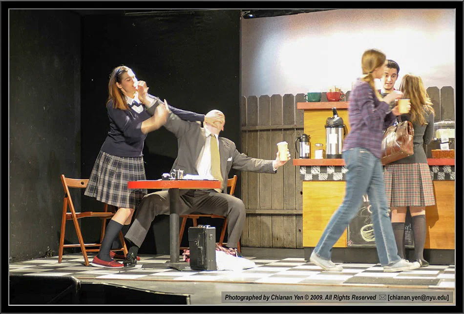 Performers converge around a seated man on a brightly lit cafe set.