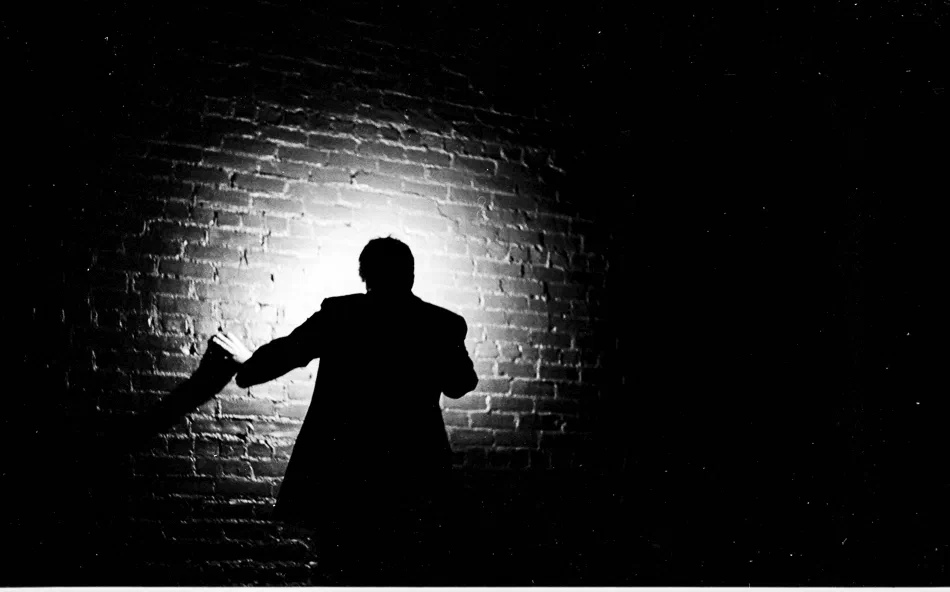 A silhouetted figure stands before a bright brick wall.