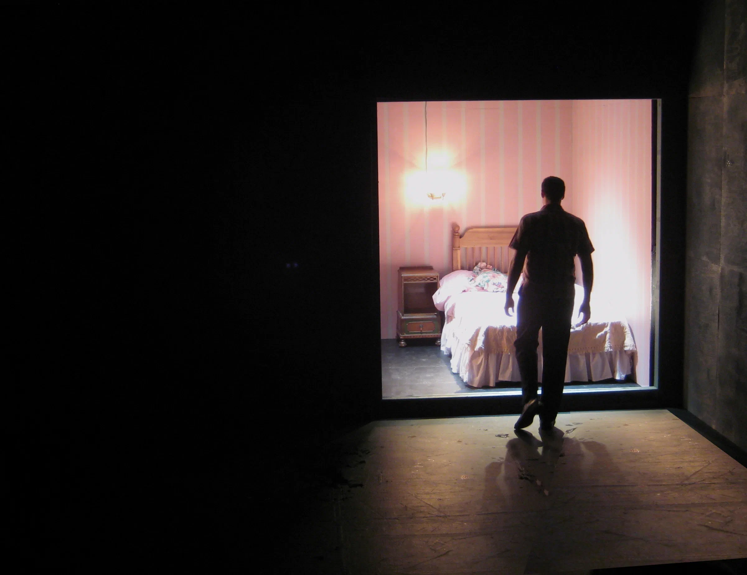A performer faces a brightly lit bedroom set framed by a dark stage.