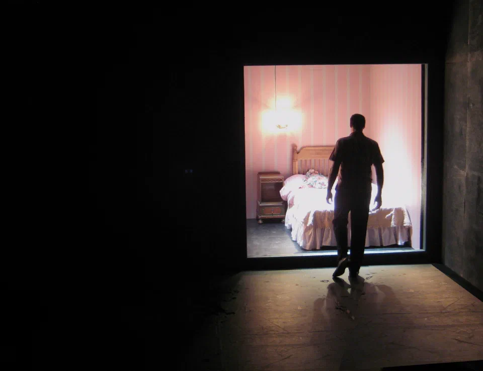 A performer faces a brightly lit bedroom set framed by a dark stage.