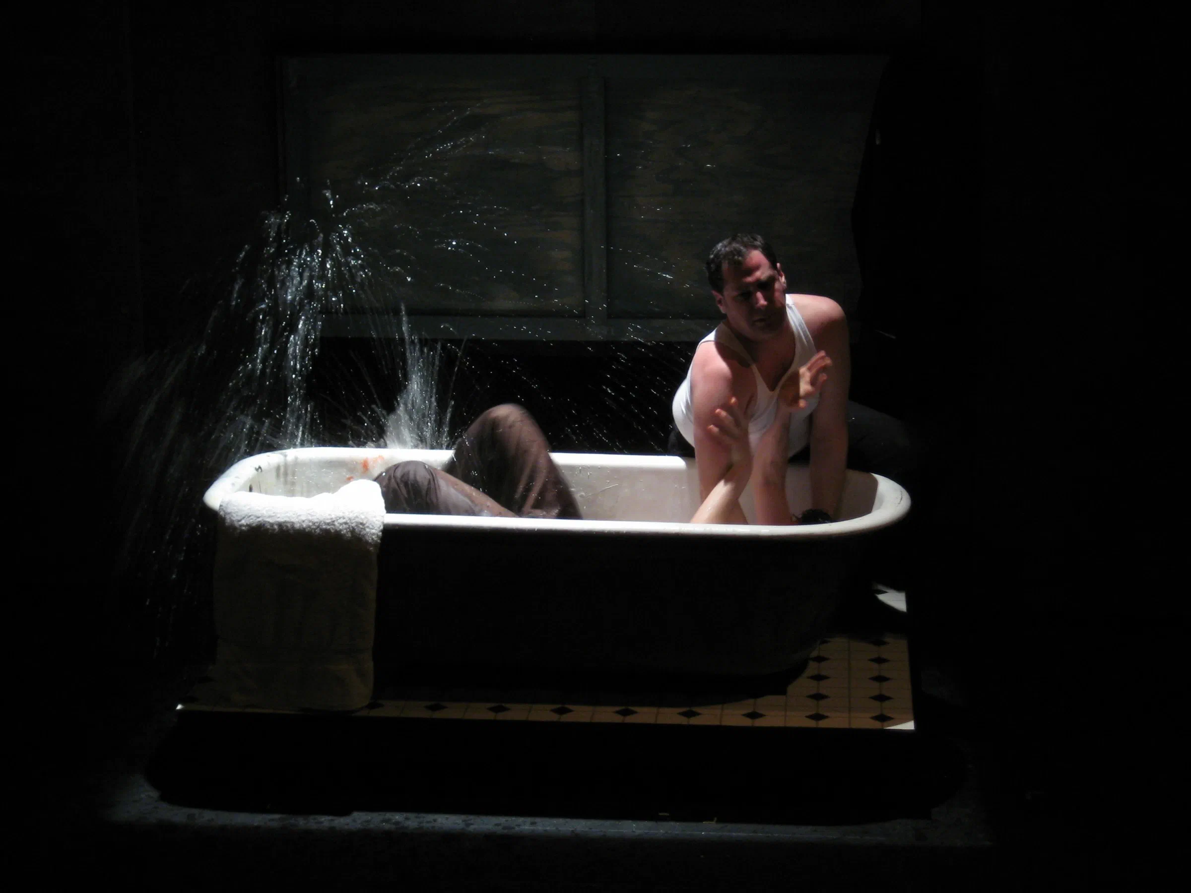 Two performers struggle in a bathtub as water sprays through the light.