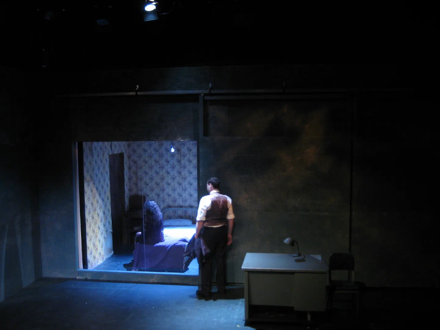 A man stands between a blue-lit bedroom and a separate writing desk.