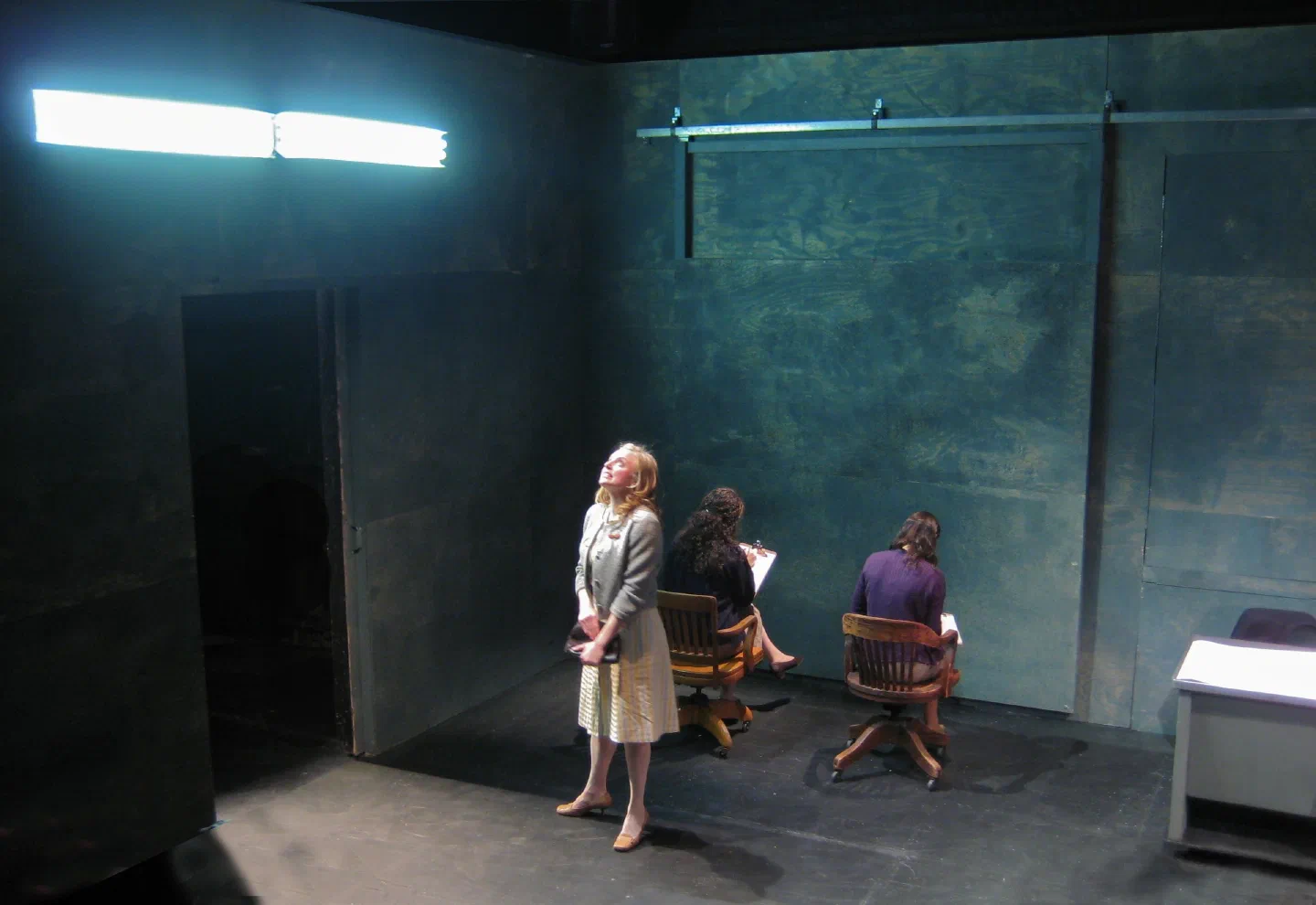 Two standing performers flank a woman lying on a bed inside a framed room.