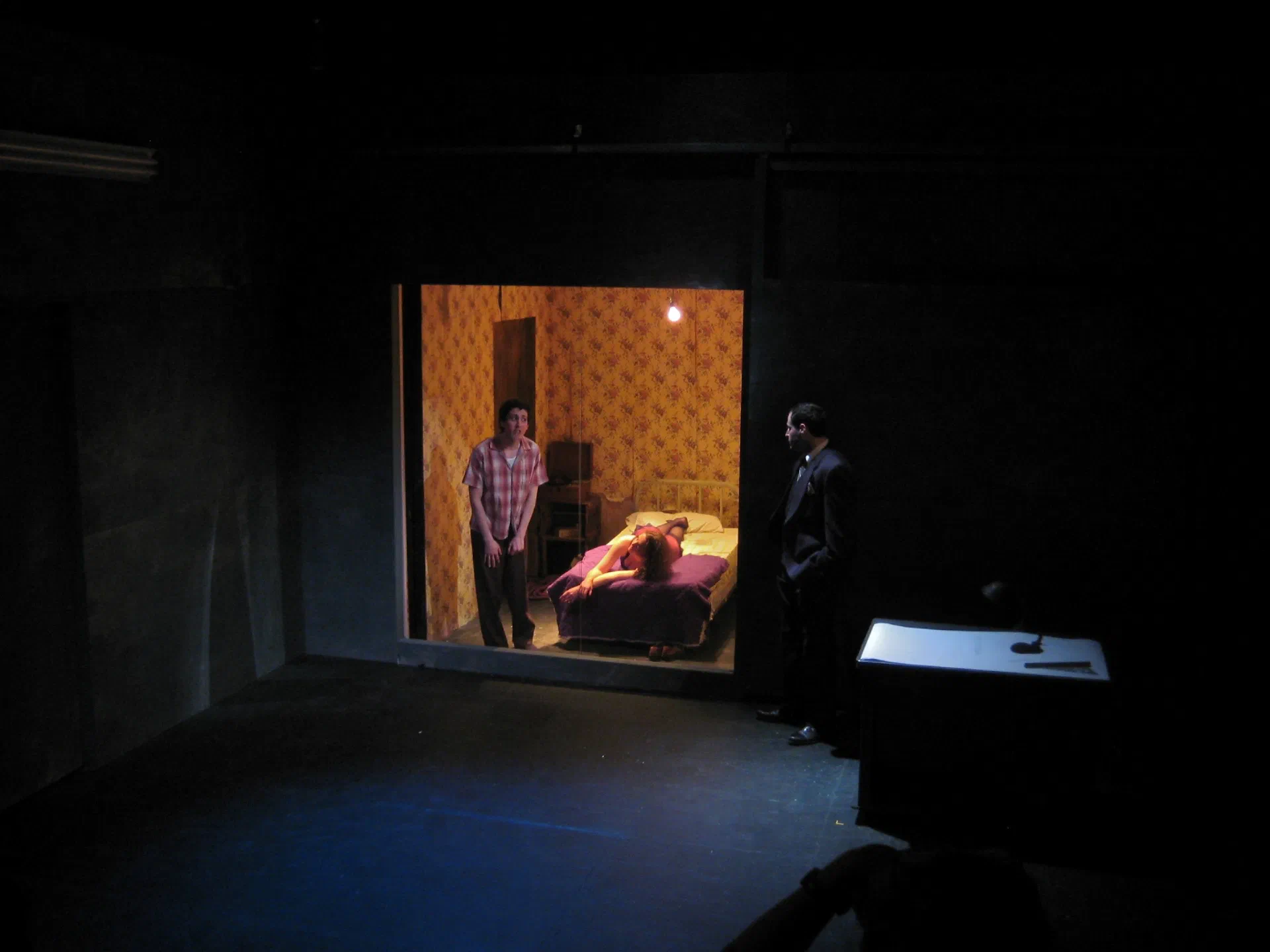 A woman stands before two seated figures at writing desks on a blue-lit stage.