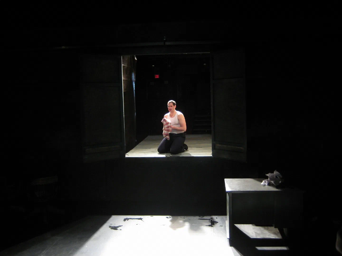 A performer kneels in a pool of light within a deep, dark stage opening.