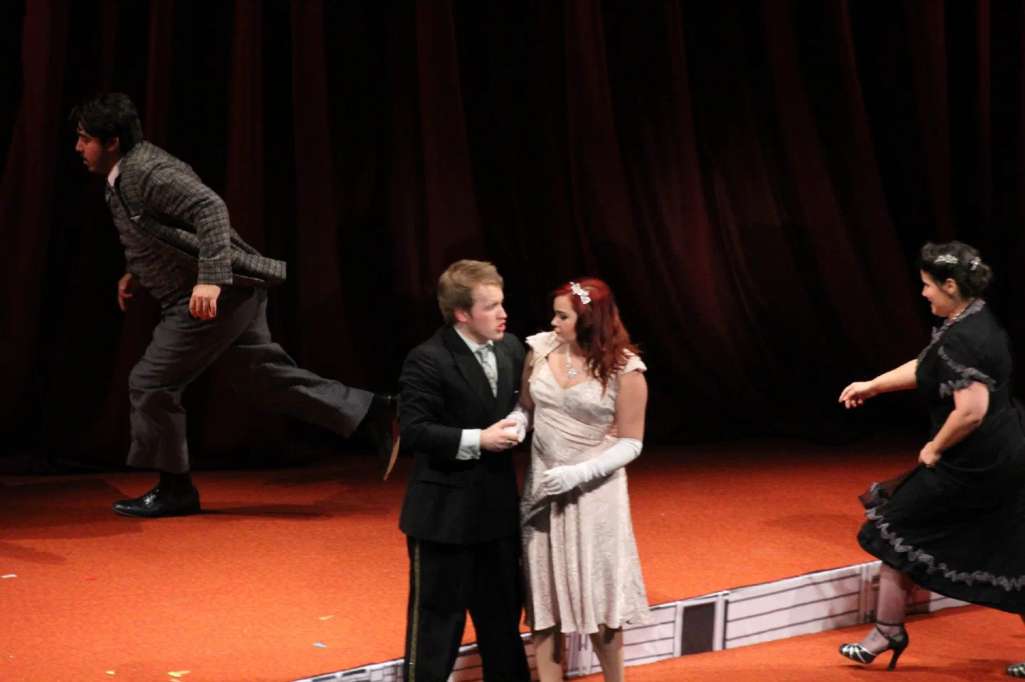 Two women approach a formally dressed couple in front of a red curtain.