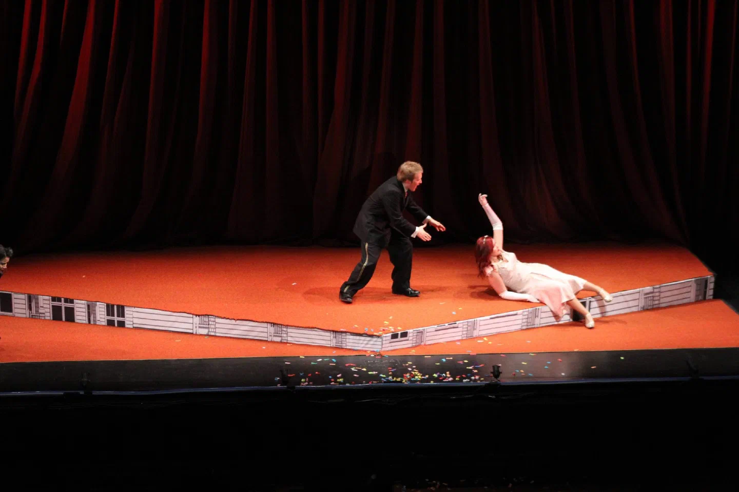 A man reaches toward a woman lying on a red platform before a dark curtain.
