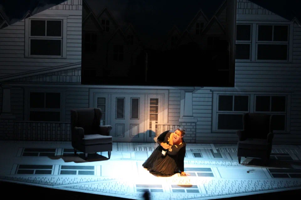 A performer kneels in a pool of light before a large house facade.