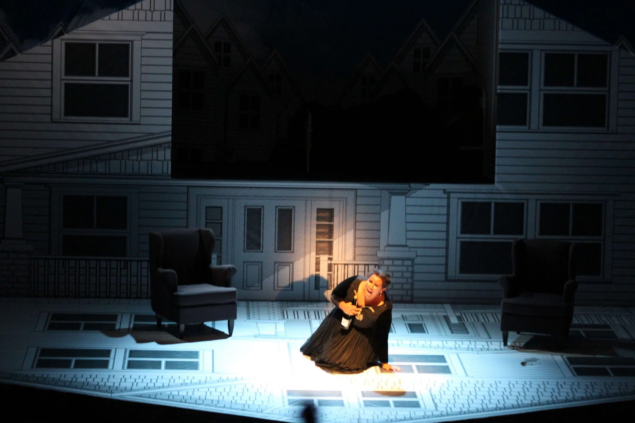 A performer kneels in a pool of light before a large house facade.