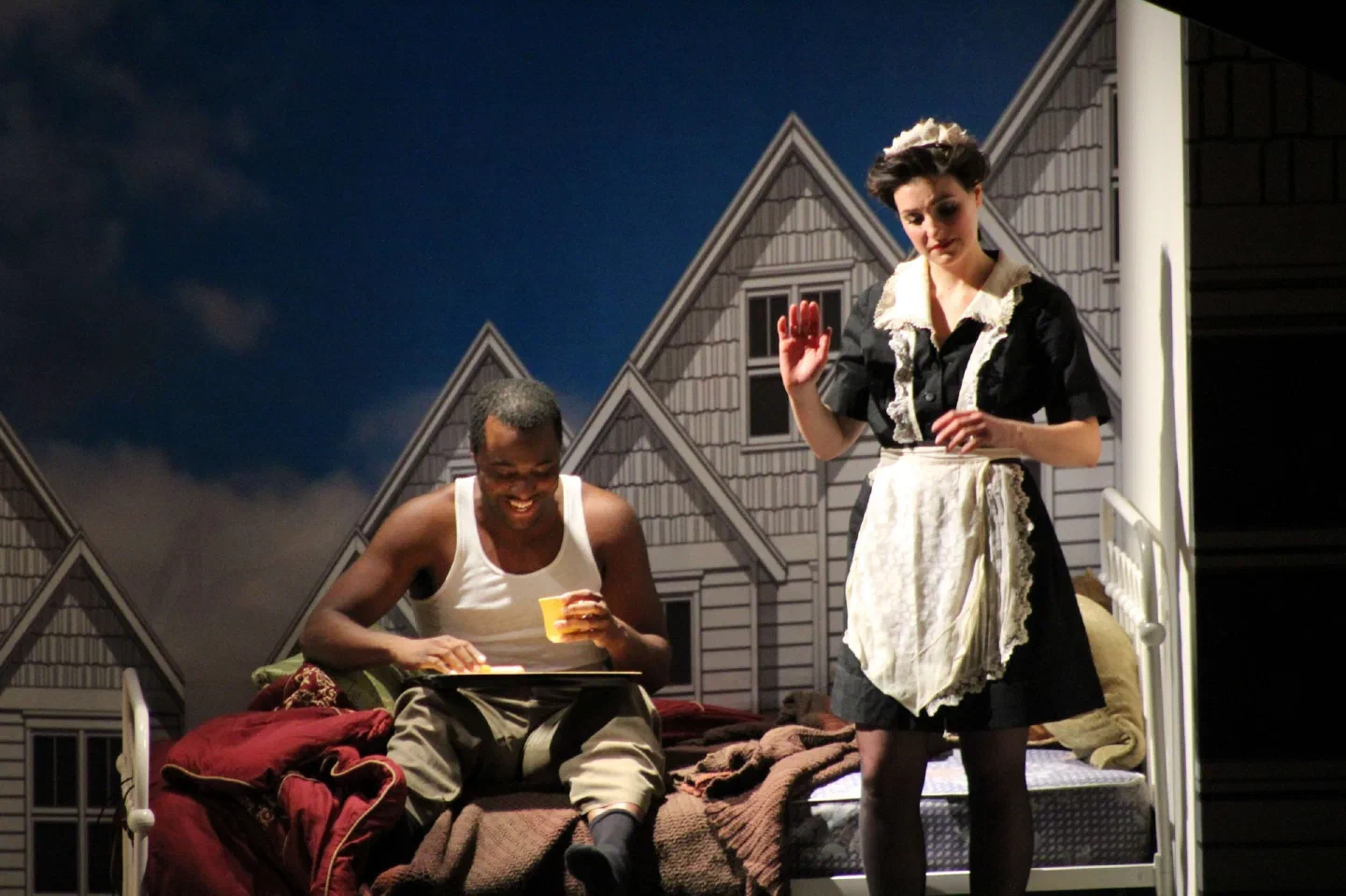 A seated man holds a drink as a maid stands beside him outside a house facade.