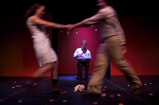 Two blurred figures circle a seated performer on a red-lit stage.