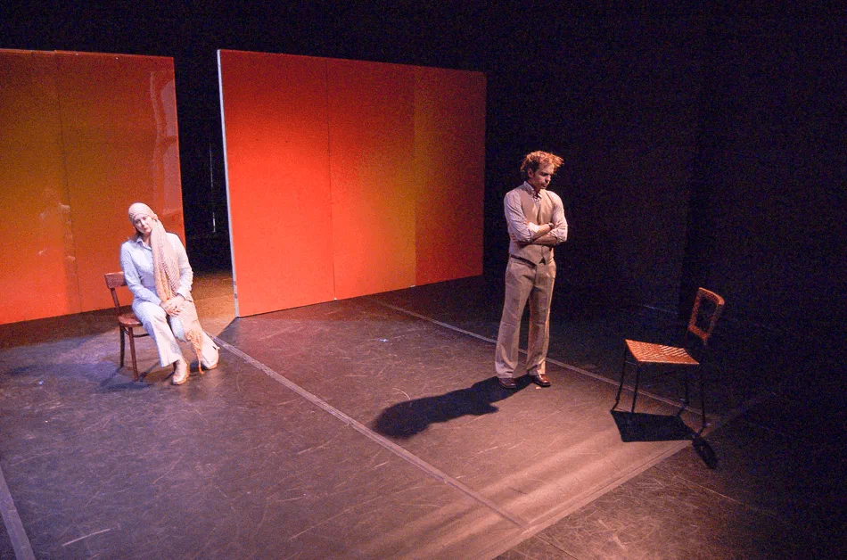 A seated man and standing woman face one another between red scenic panels.