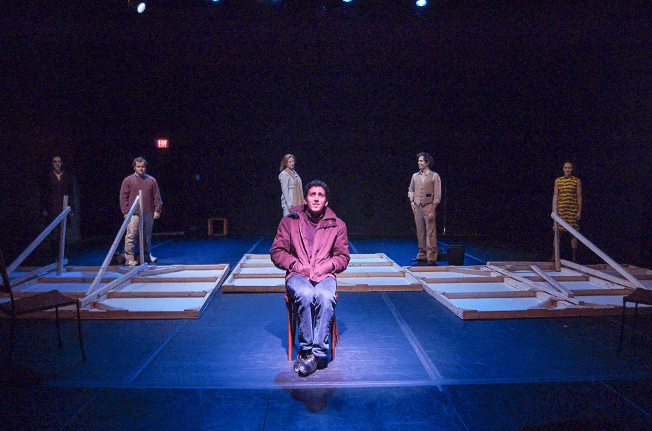 An ensemble stands across separated platforms while a seated man faces the audience.