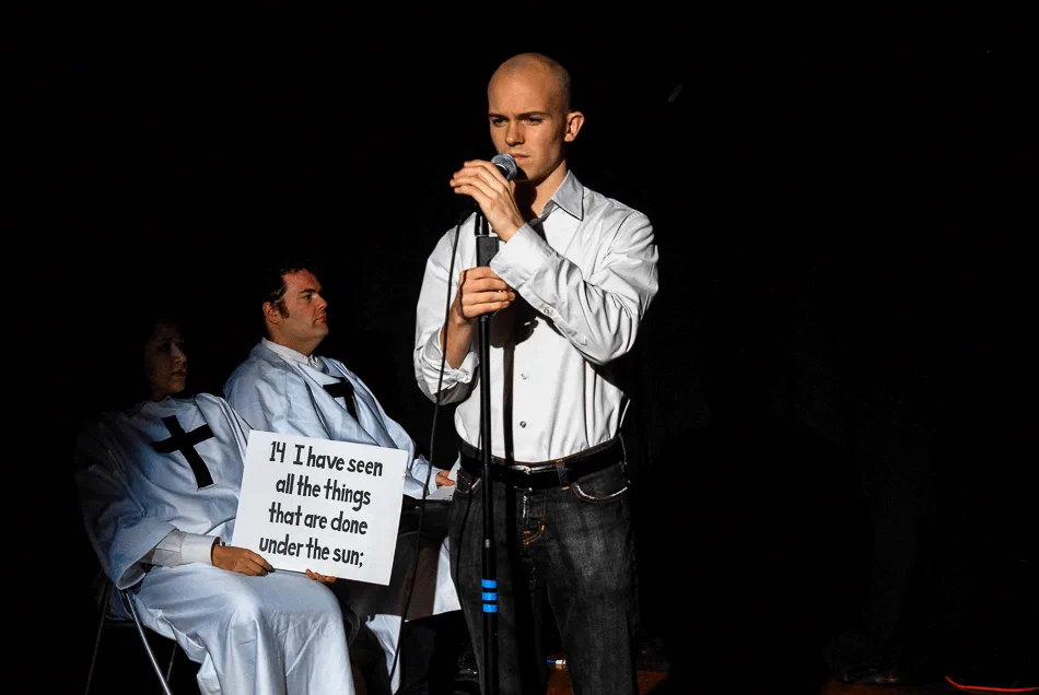 A man speaks into a microphone beside a seated performer in a white robe holding a sign.