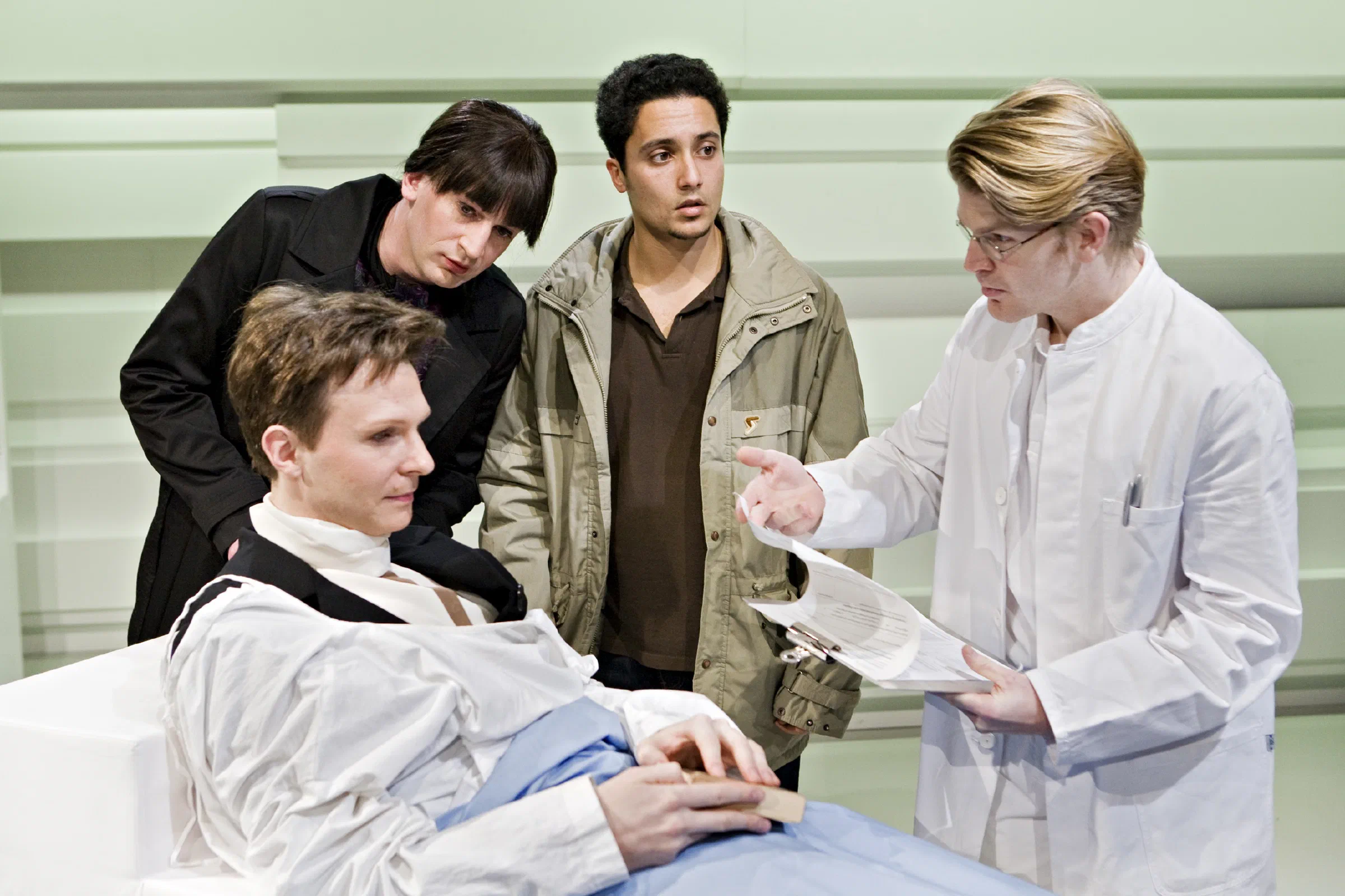 Four performers cluster around a seated figure on a pale clinical set.