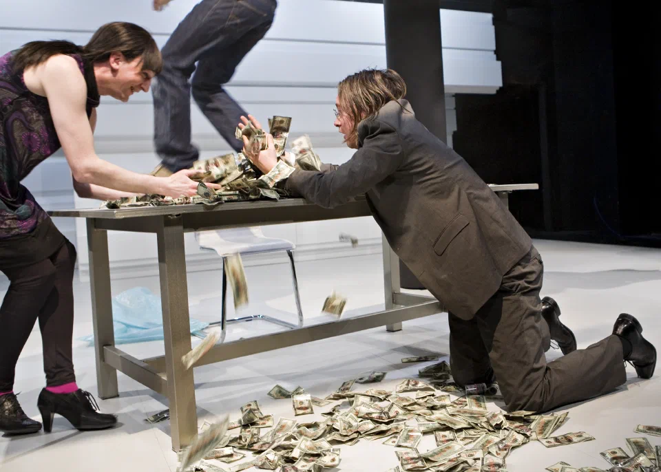 Performers struggle across a table scattered with paper money.