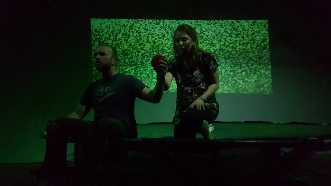 Two performers sit before a rectangular field of green projected static.