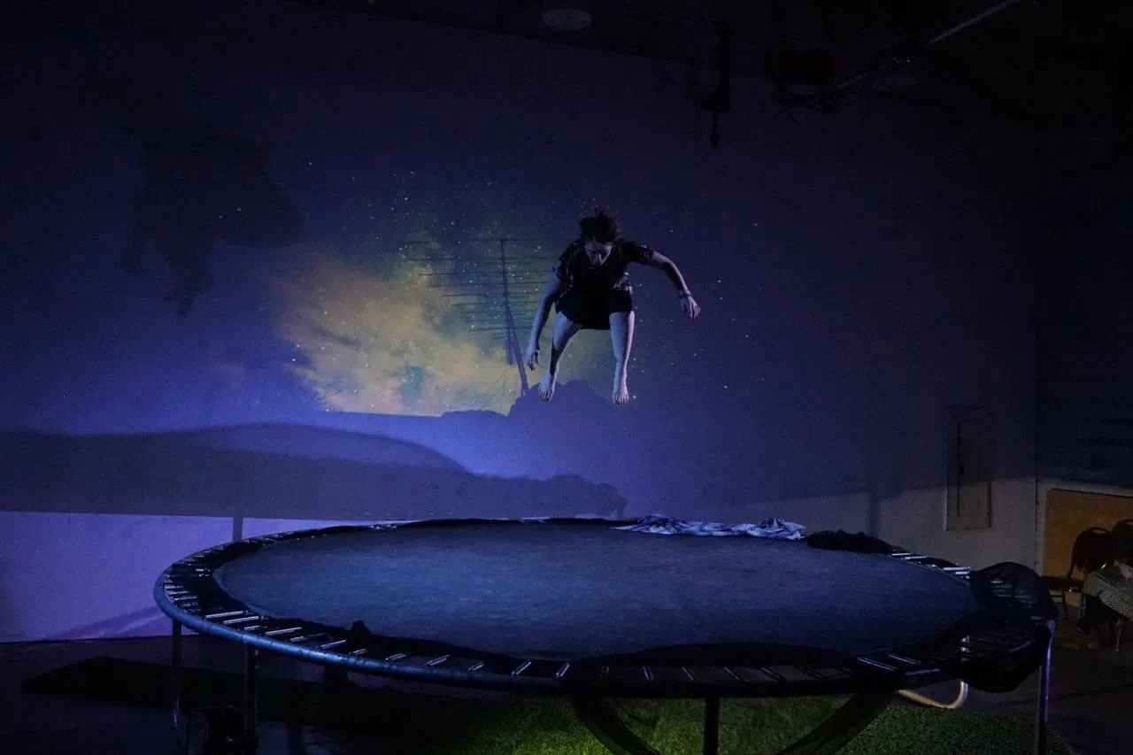 A performer crouches in midair above a trampoline beneath a landscape projection.