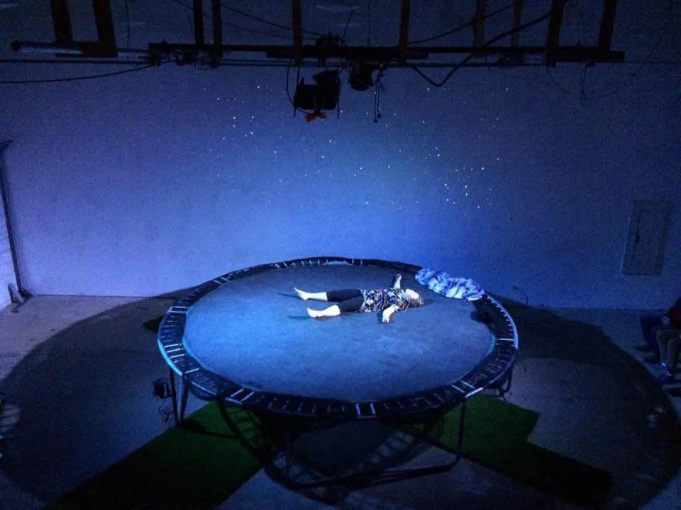 A performer lies at the center of a trampoline under cool blue light.