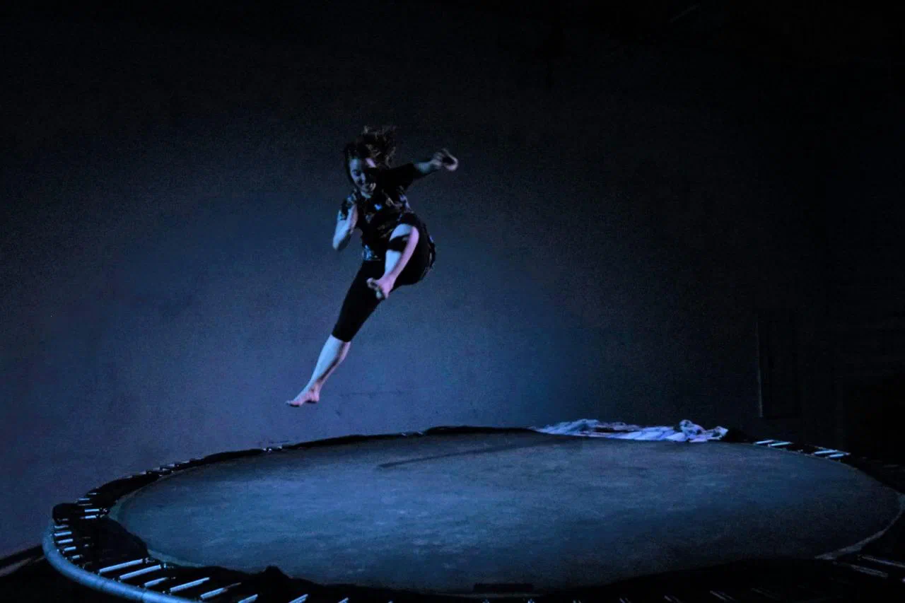 A performer hangs in midair above a trampoline on a dark stage.