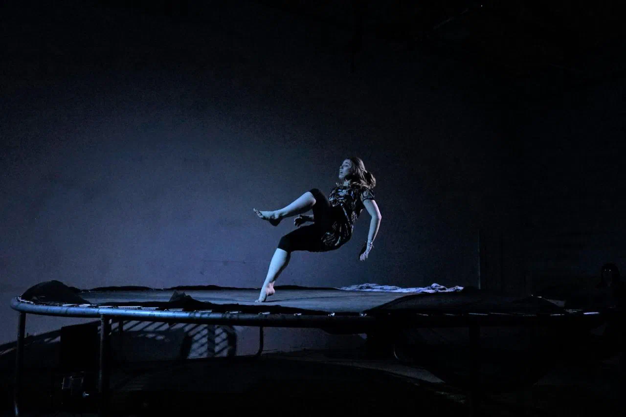 A performer falls backward toward a trampoline under blue stage light.