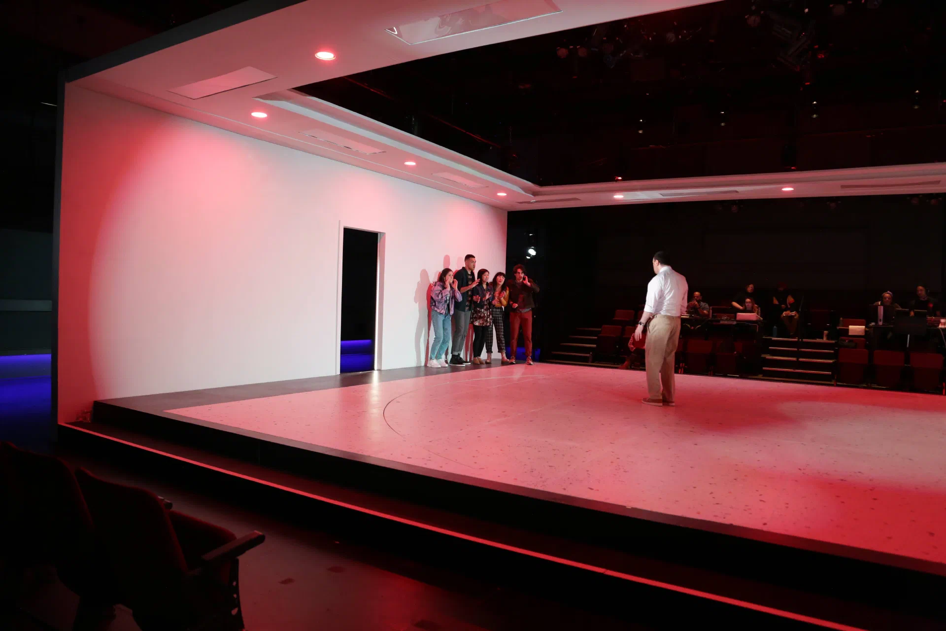 A performer faces an ensemble gathered along the side of a red-lit white stage.