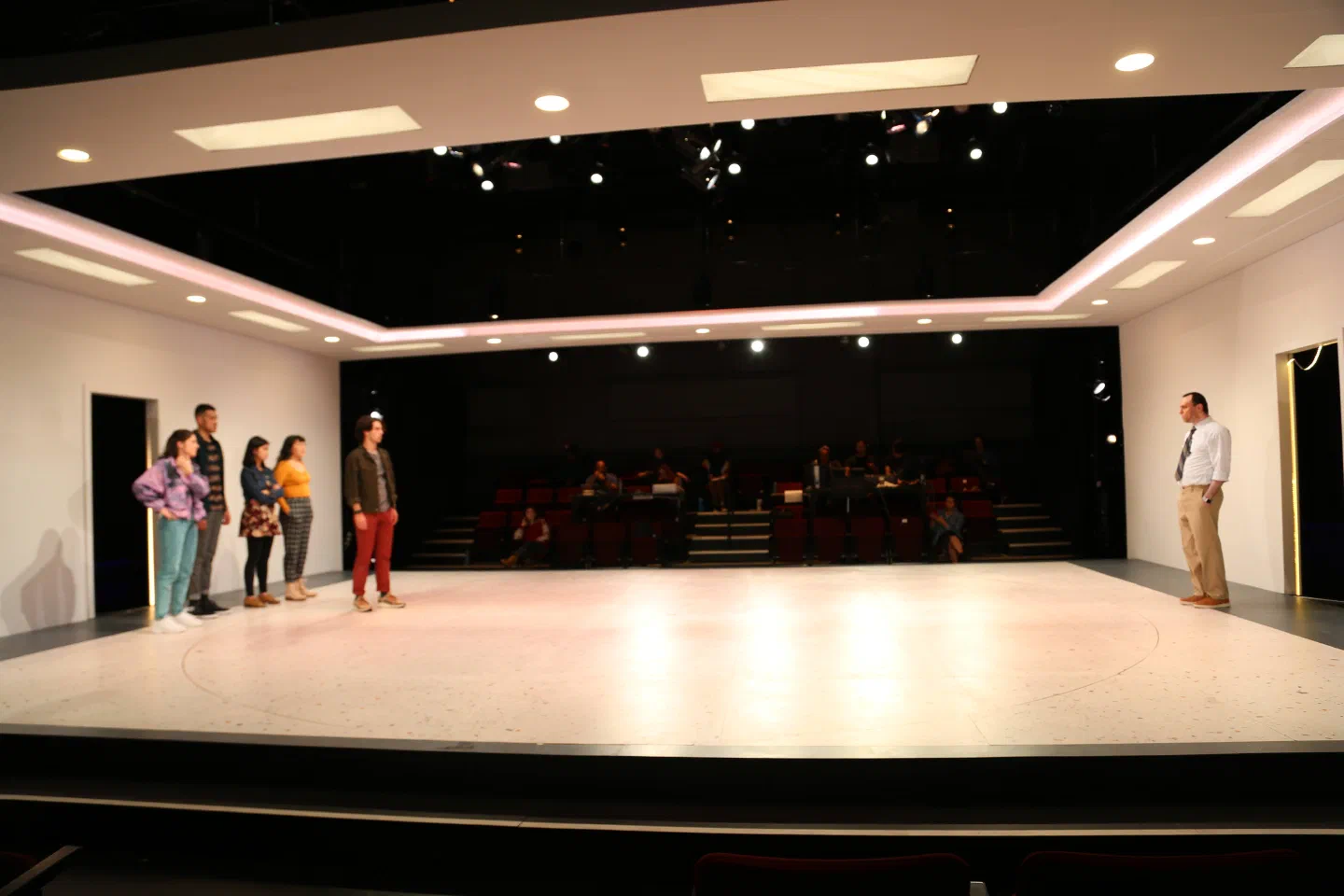 Performers stand at opposite sides of a broad white stage framed by glowing ceiling panels.