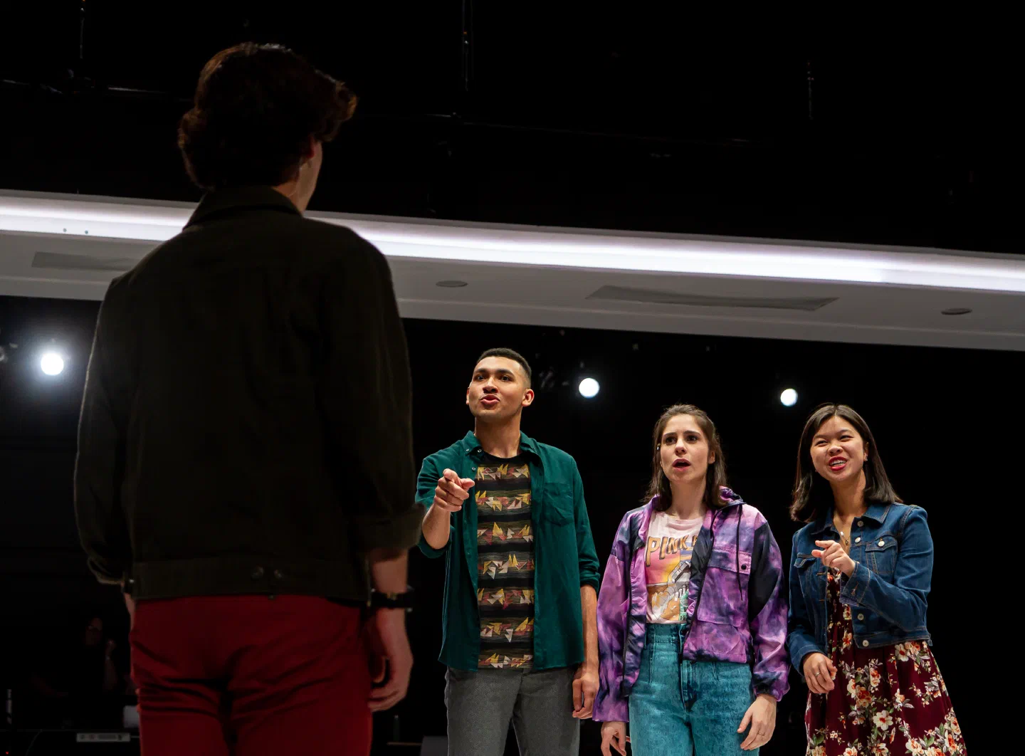 Three performers address another figure on a dark stage beneath bright overhead lights.