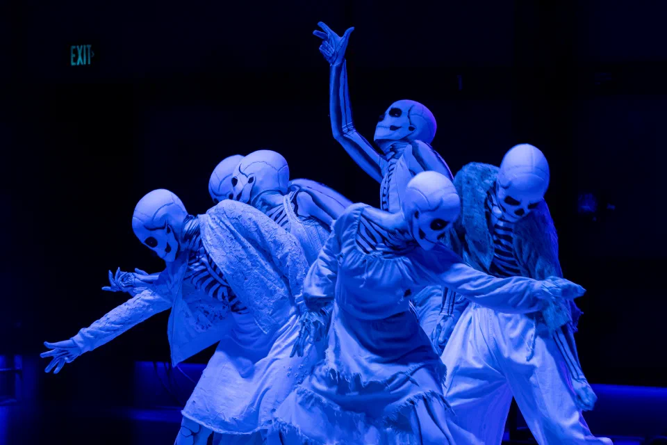 Five performers in pale skeletal costumes reach outward under blue light.