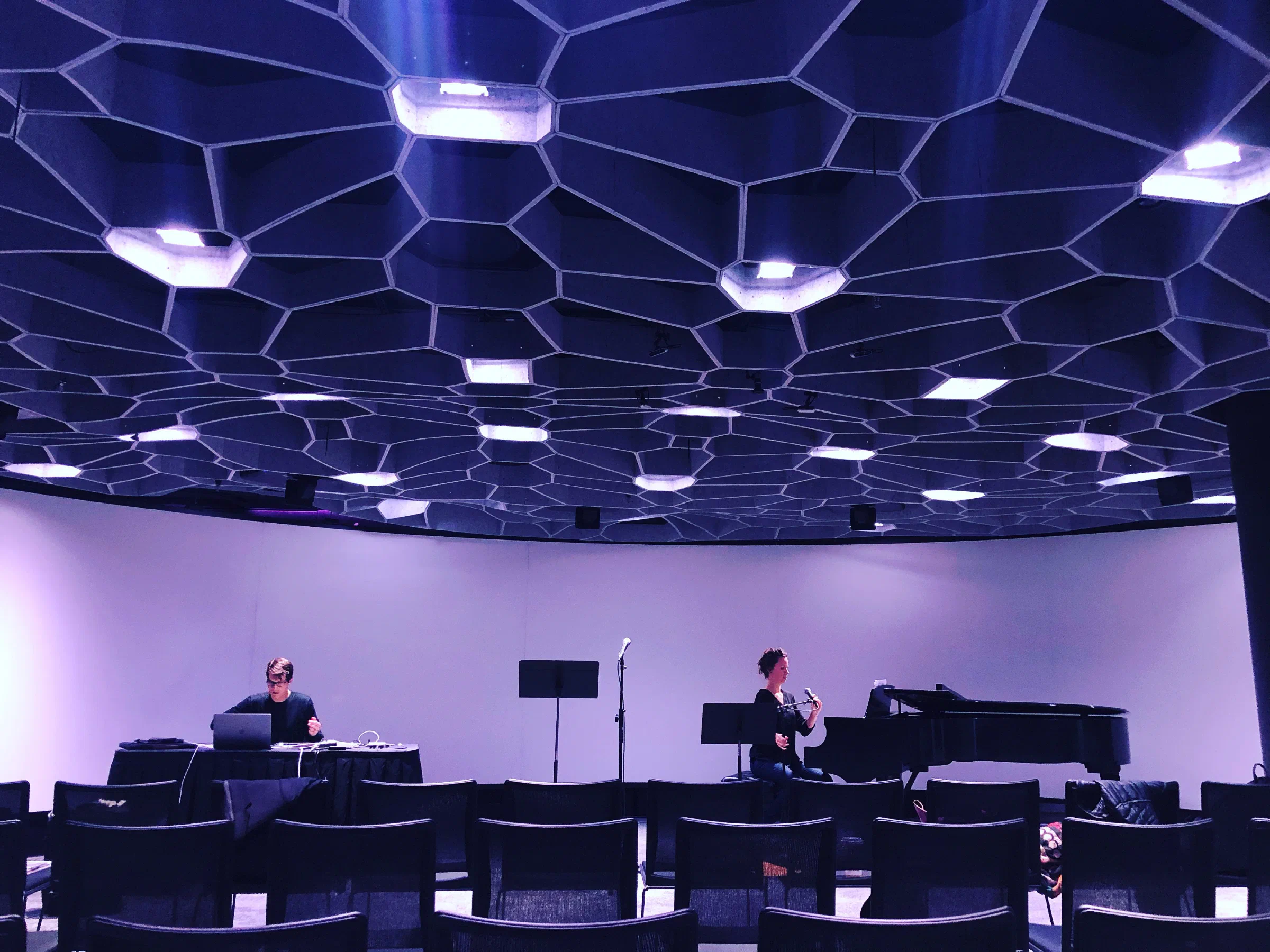 A pianist and performer rehearse beneath a geometric concert-hall ceiling.