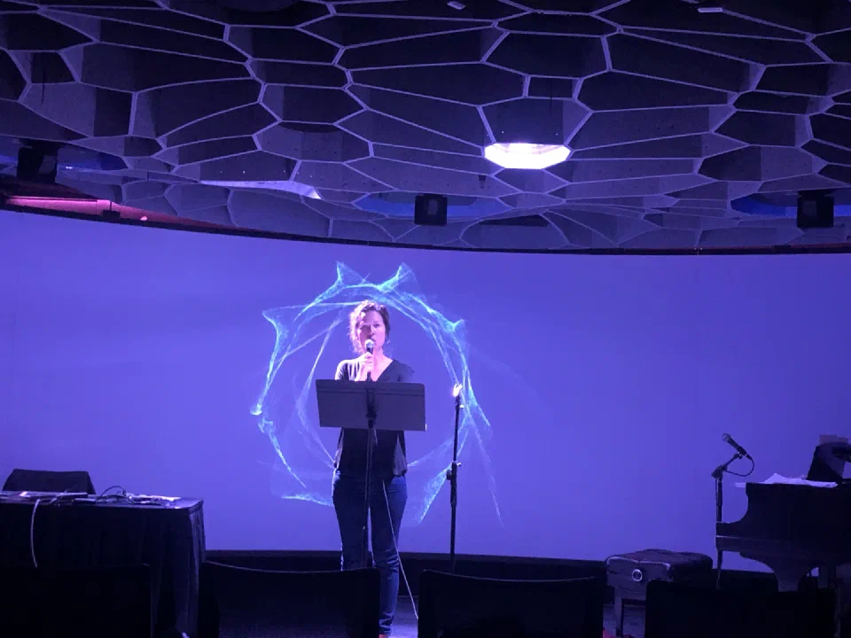 A performer stands before a luminous circular projection in a concert hall.