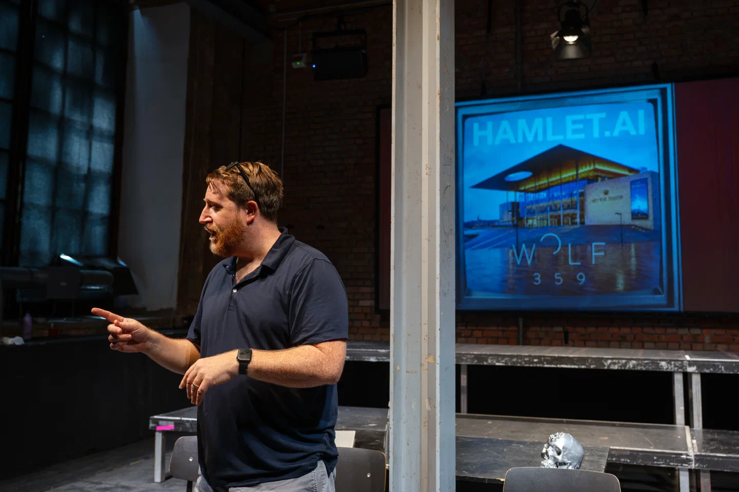 A man gestures from behind a clear scenic panel with a Hamlet.ai projection behind him.