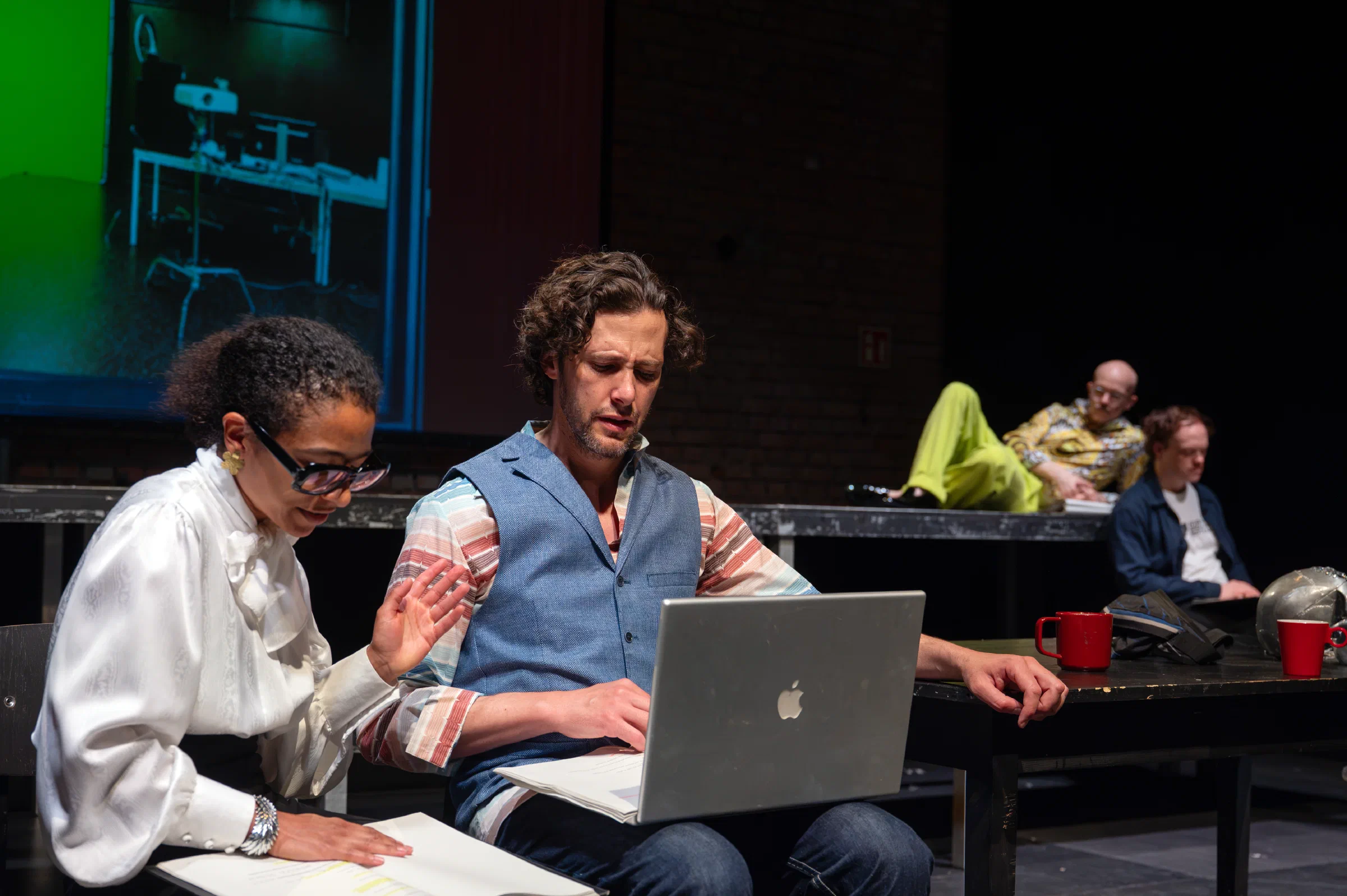 Performers work at laptops while two figures sit on a raised platform during Hamlet.ai.