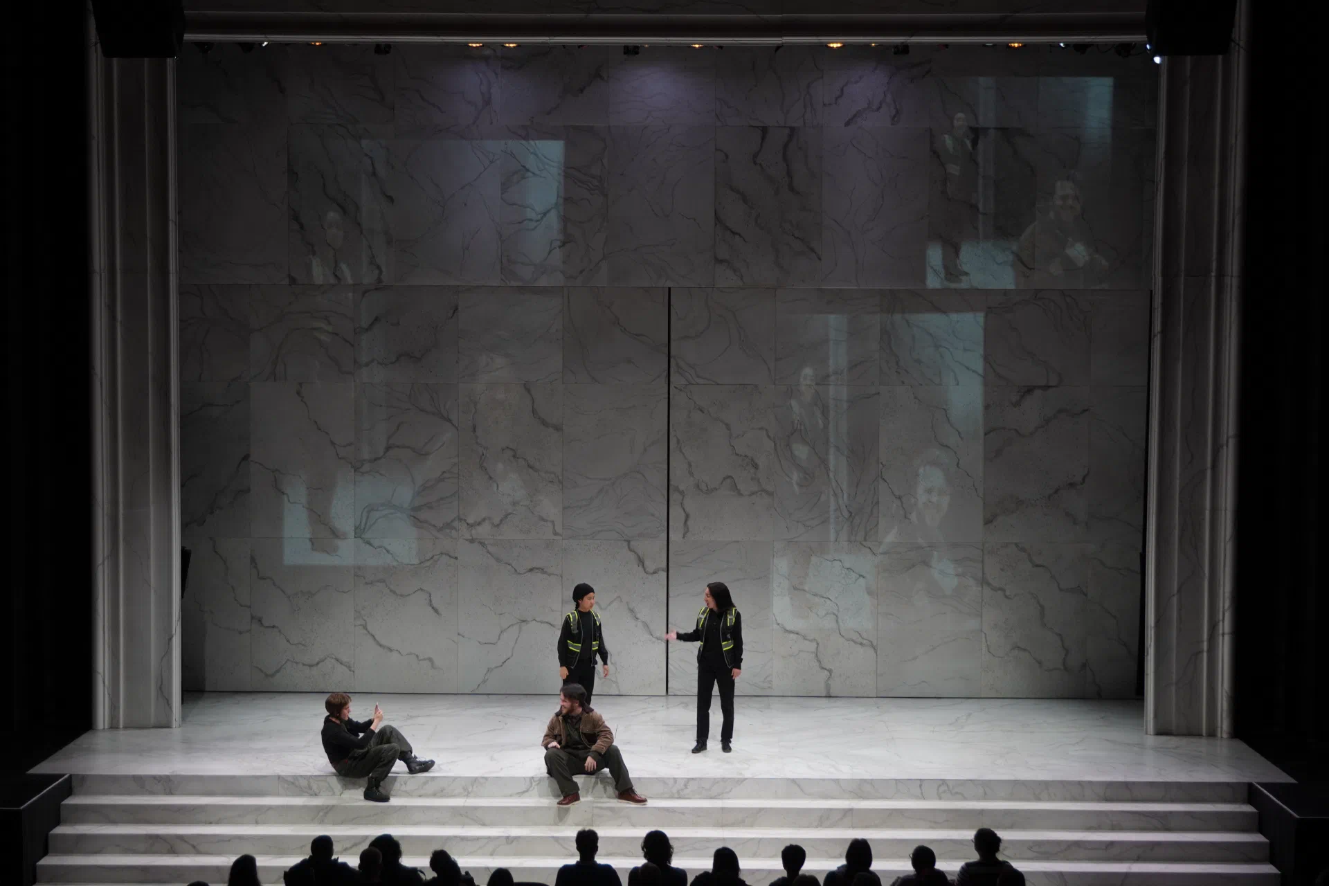 Performers occupy a broad pale stone set as audience members watch from below the stage.