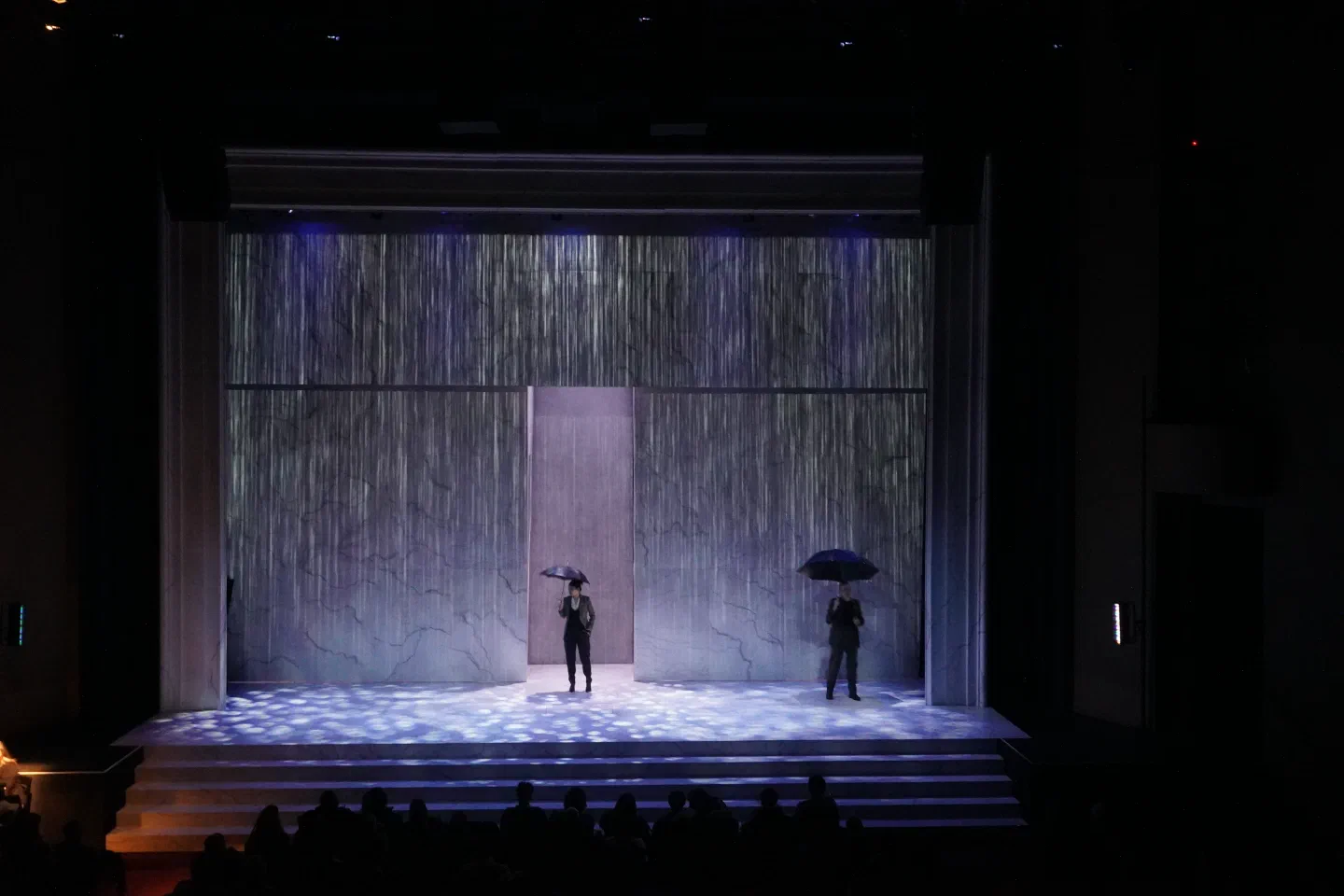 Two performers carrying umbrellas stand beneath falling rain within a tall doorway.
