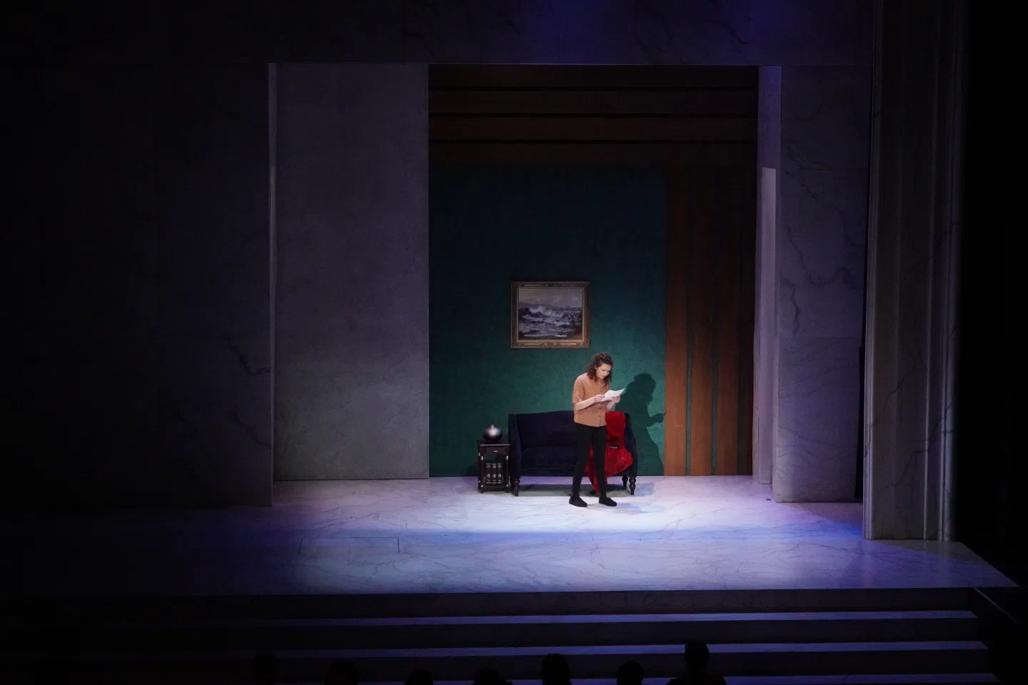 Two performers sit together beside a red chair in a recessed room.
