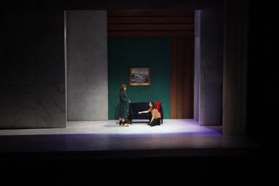 One performer stands over another beside a red chair in a tall recessed room.