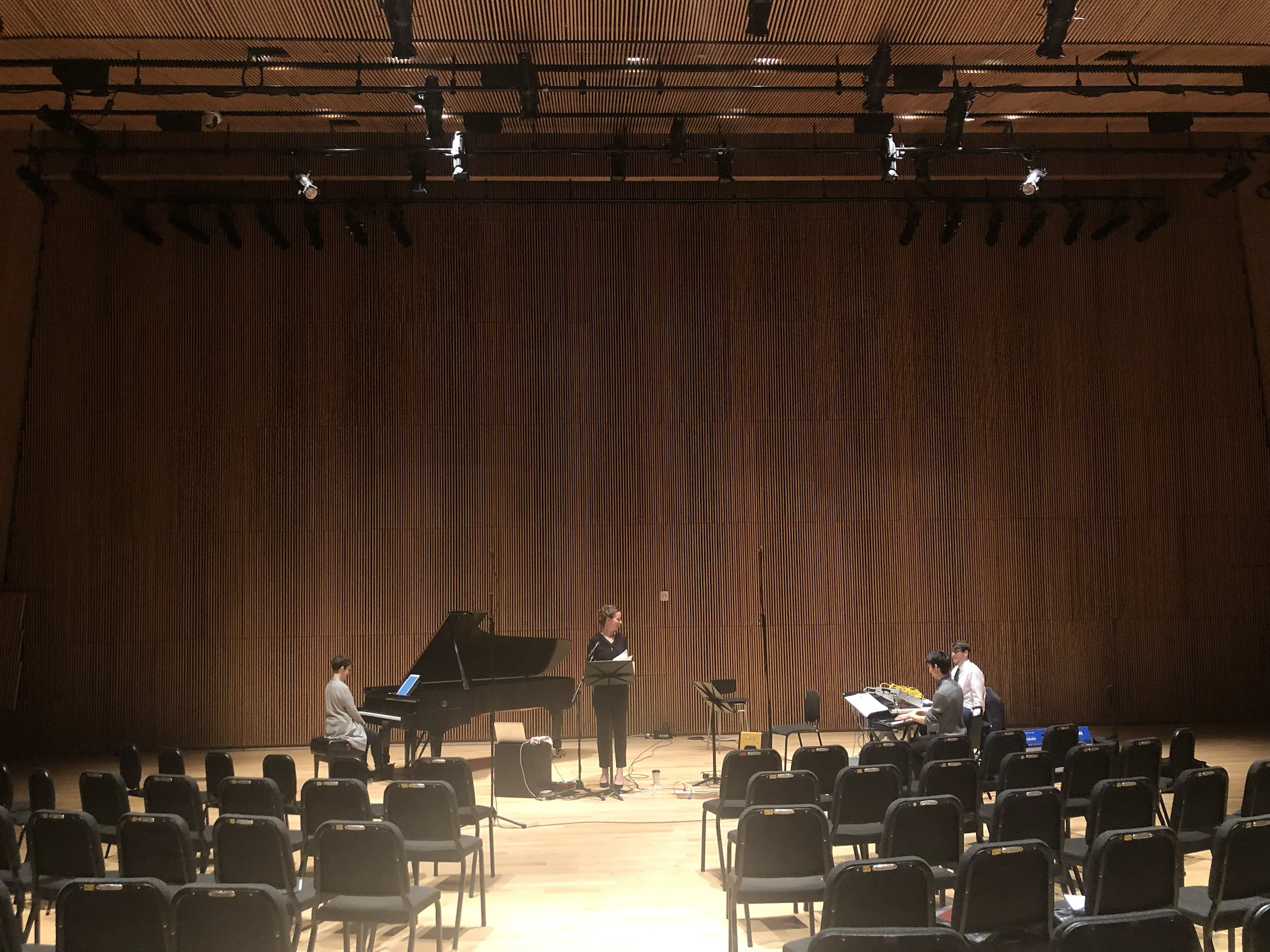 Musicians perform on a wood-paneled concert stage viewed from the audience.