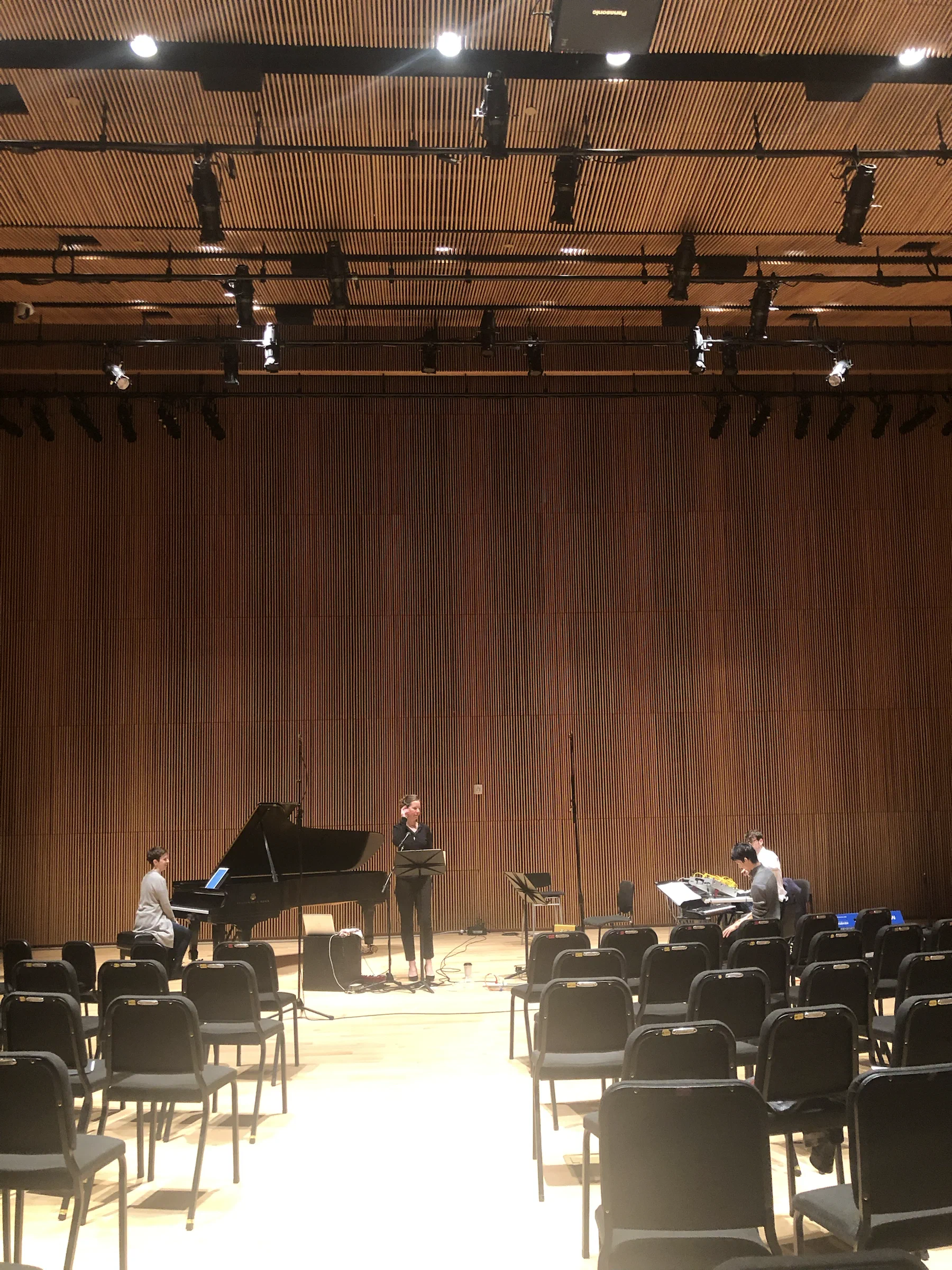 Musicians rehearse around a grand piano on a wood-paneled concert stage.