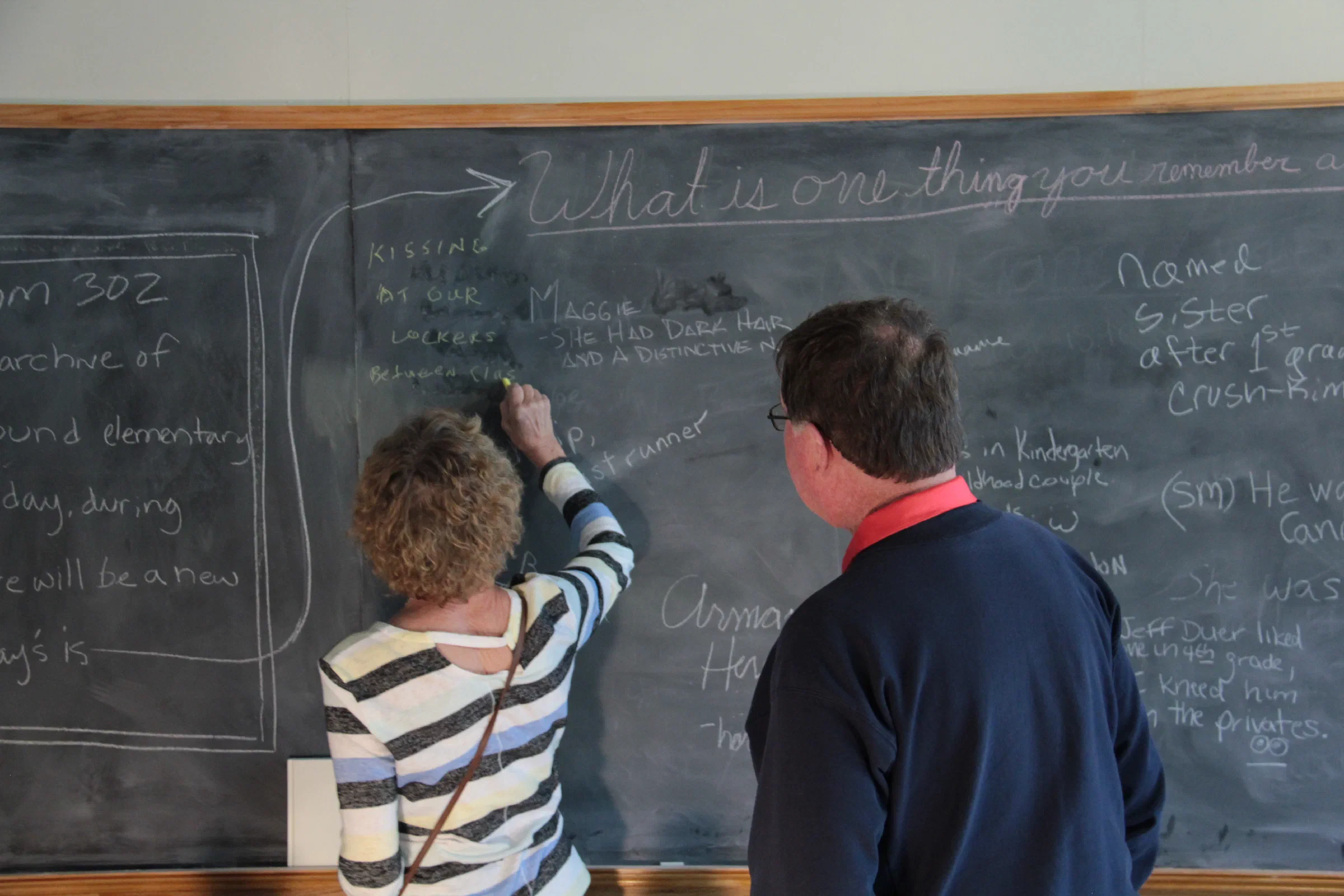 Two visitors write on a chalkboard filled with memories and handwritten notes.
