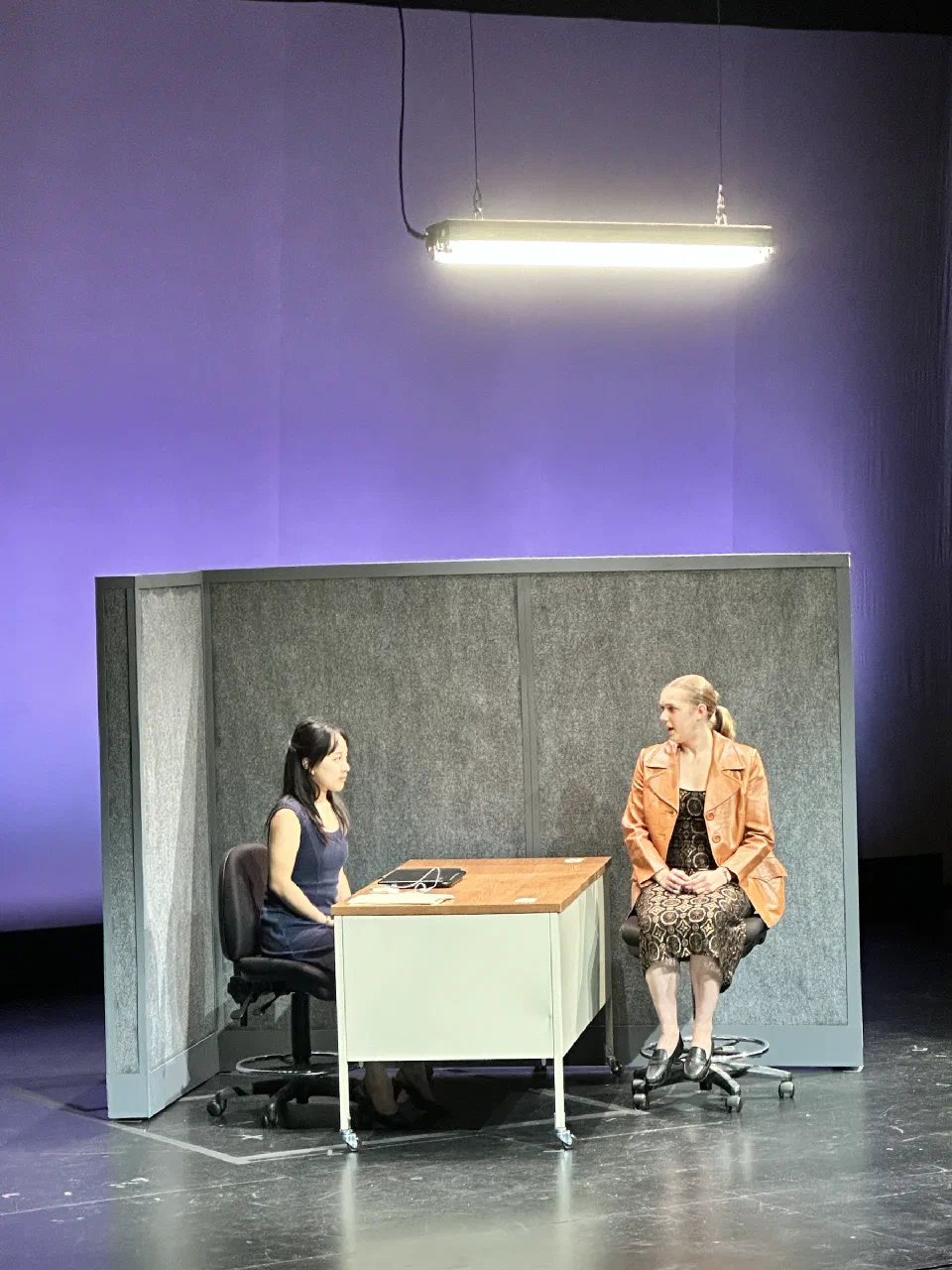 Two performers sit across a small table beneath a suspended fluorescent light.