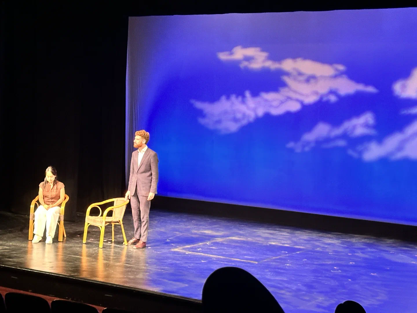Two performers sit apart on a blue stage beneath a small chandelier.